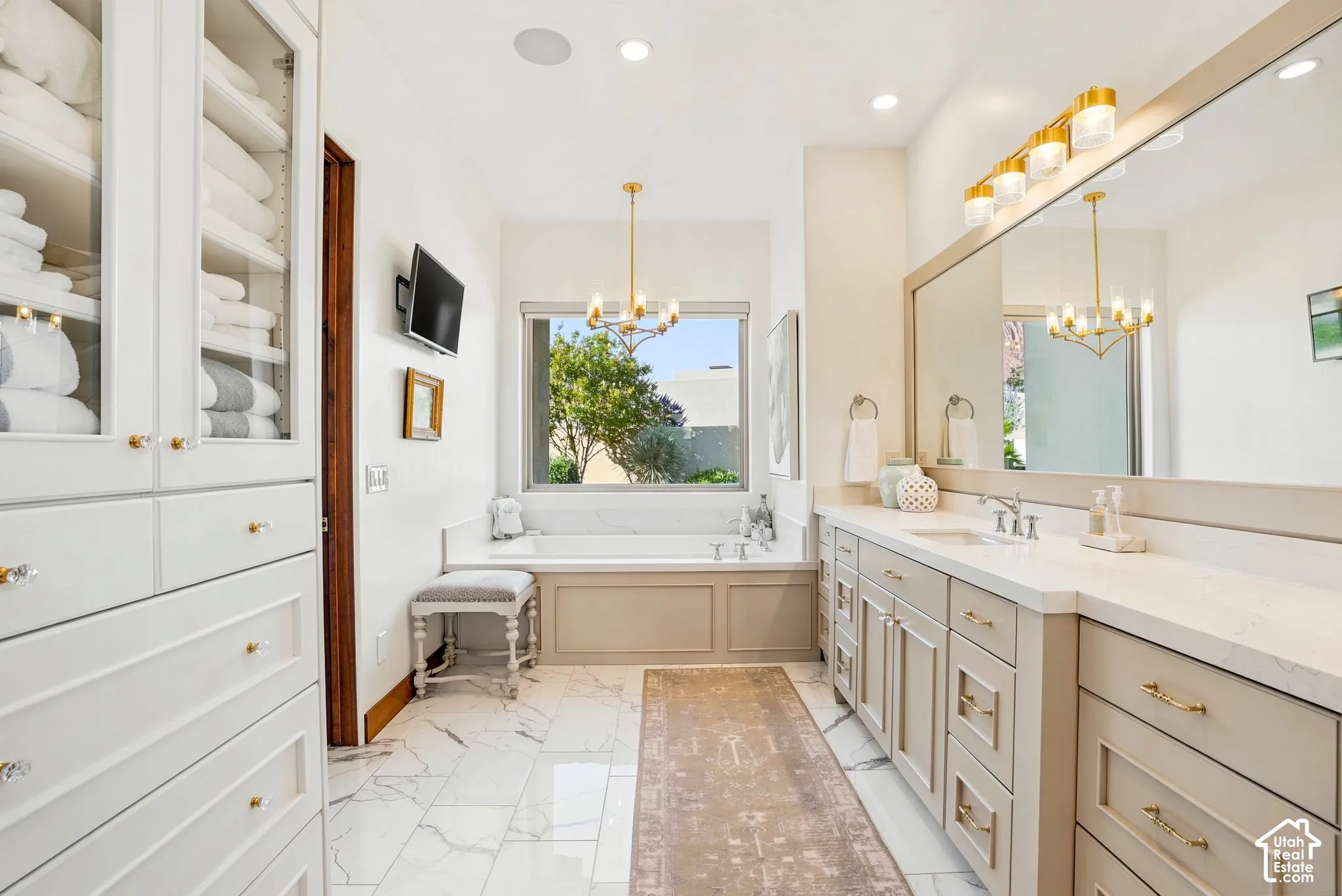 Built-in white cabinetry with glass fronts, recessed bathtub with a window, dual vanity with undermount sinks and light countertops, large wall-mounted mirror, and white tile flooring