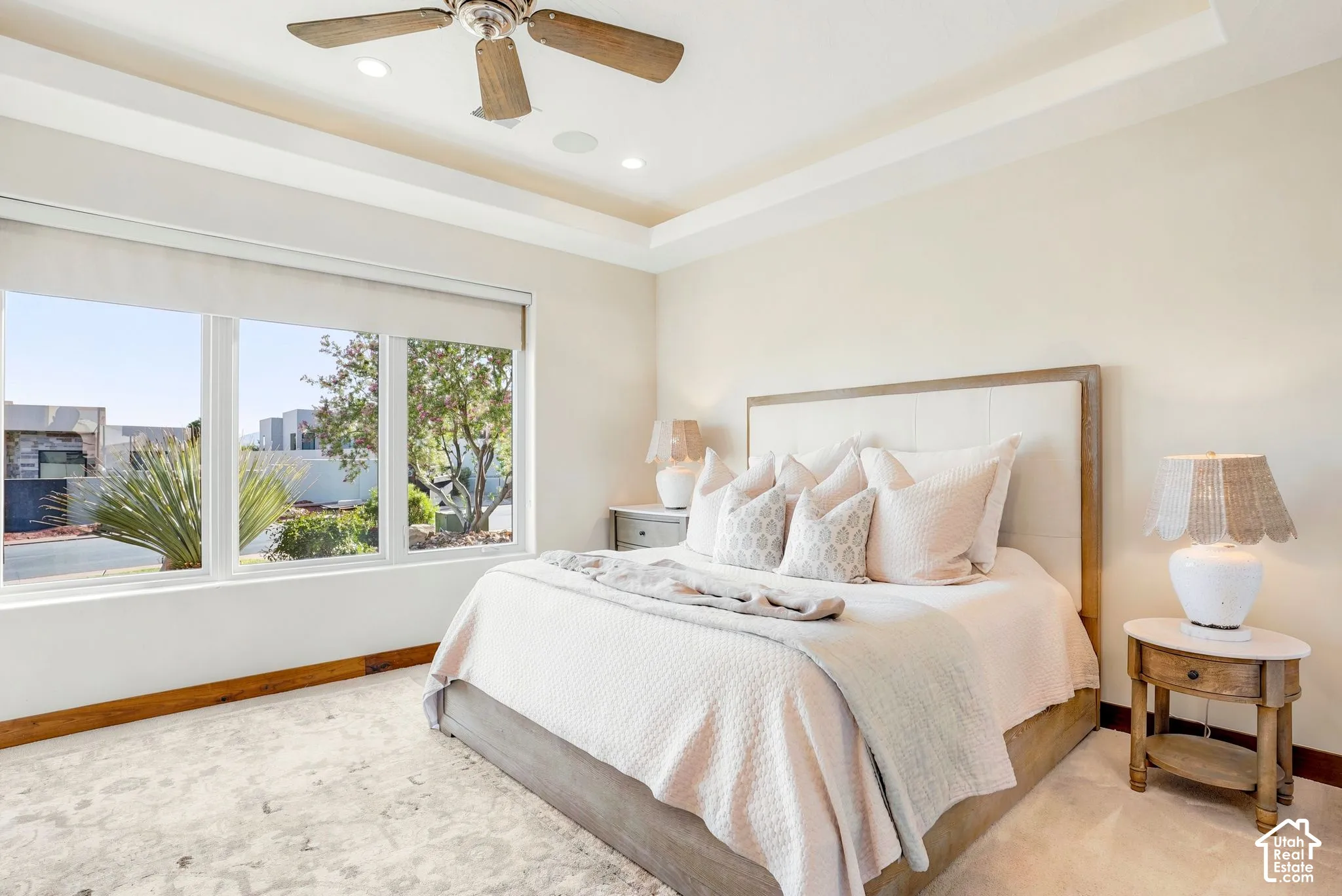Tray ceiling with recessed lighting, ceiling fan with wood-finish blades, large three-pane window, upholstered headboard, and wood-finish trim along the wall