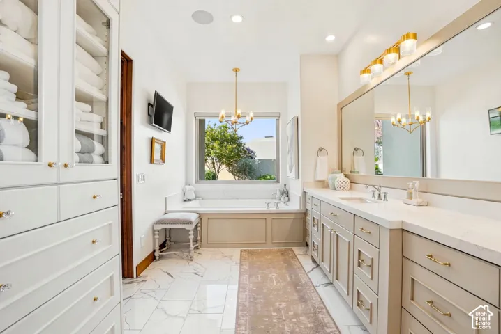 Built-in white cabinetry with glass fronts, recessed bathtub with a window, dual vanity with undermount sinks and light countertops, large wall-mounted mirror, and white tile flooring