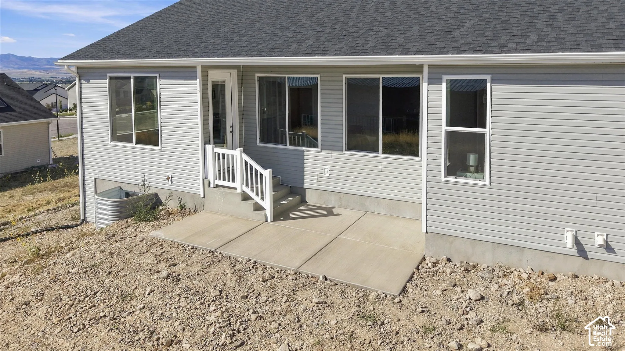 Light gray siding exterior with a dark shingle roof, featuring a concrete patio, white railing, and a glass-paneled door