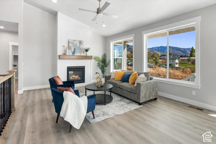 Vaulted ceiling with a ceiling fan, fireplace with a wood mantle, wood-finish flooring, and large windows with mountain scenery