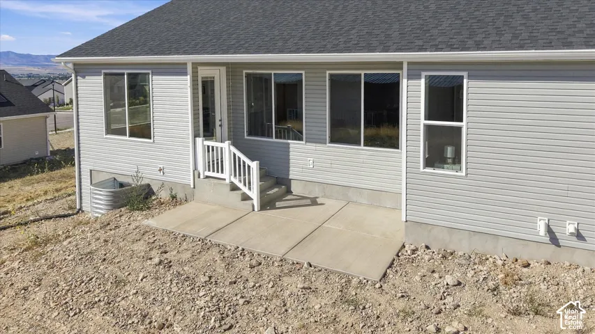 Light gray siding exterior with a dark shingle roof, featuring a concrete patio, white railing, and a glass-paneled door