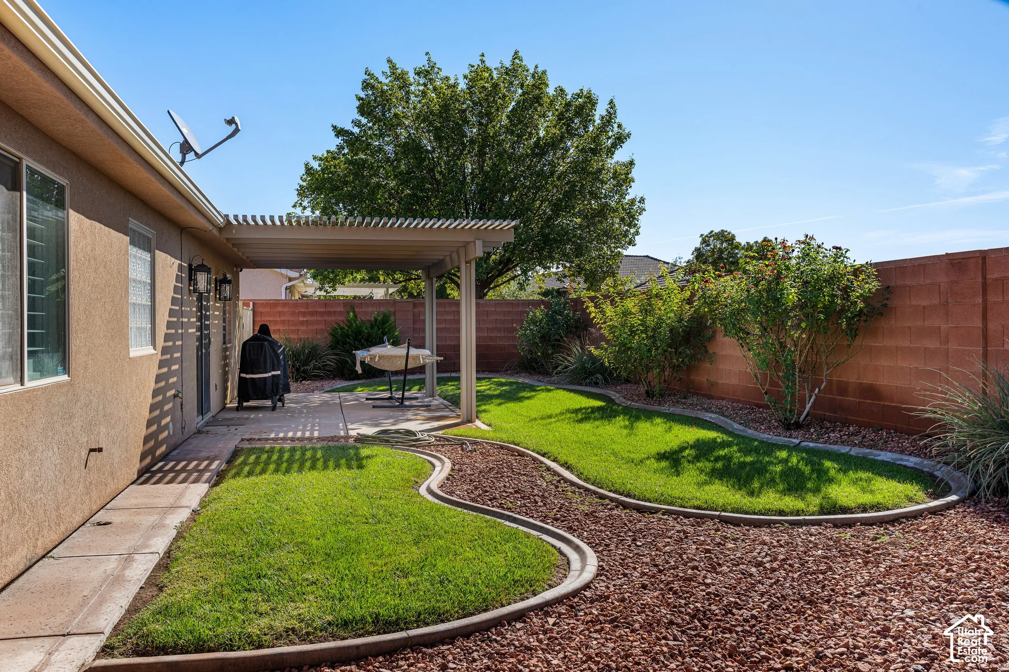 Rear yard featuring a covered patio, concrete paver walkway, landscaped beds with gravel and mature shrubs, and a block wall perimeter