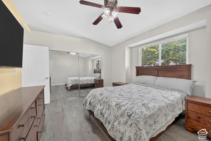Vaulted ceiling with a ceiling fan light fixture, wood-finish flooring, mirrored closet doors, and a large window with blinds