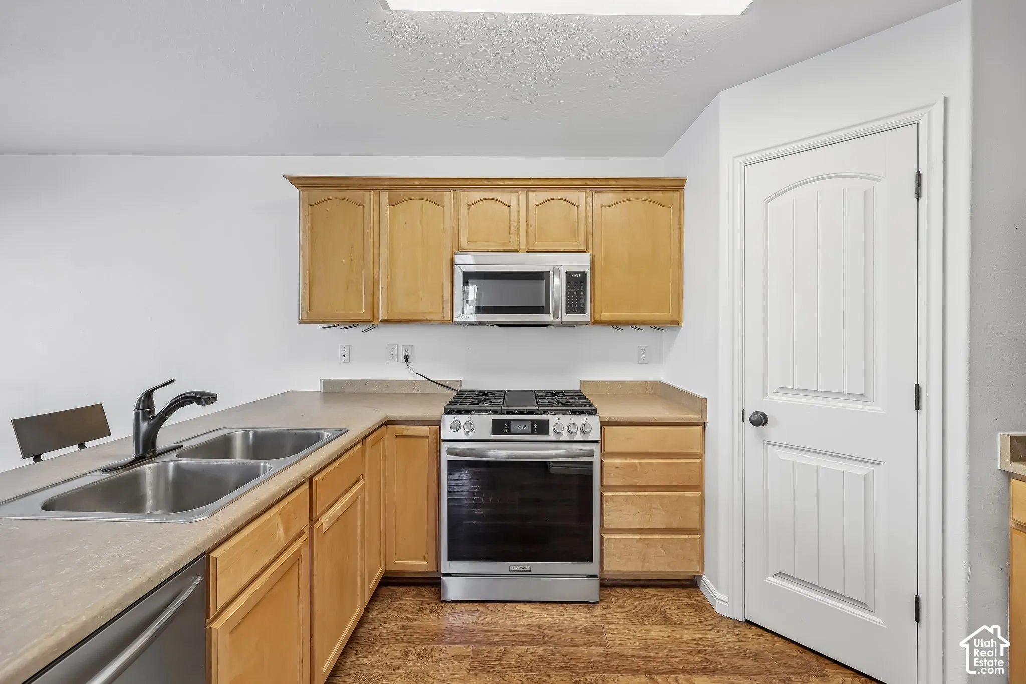 Wood-finish flooring, light wood cabinetry, stainless steel appliances, double basin sink with a matte black faucet, and a paneled interior door
