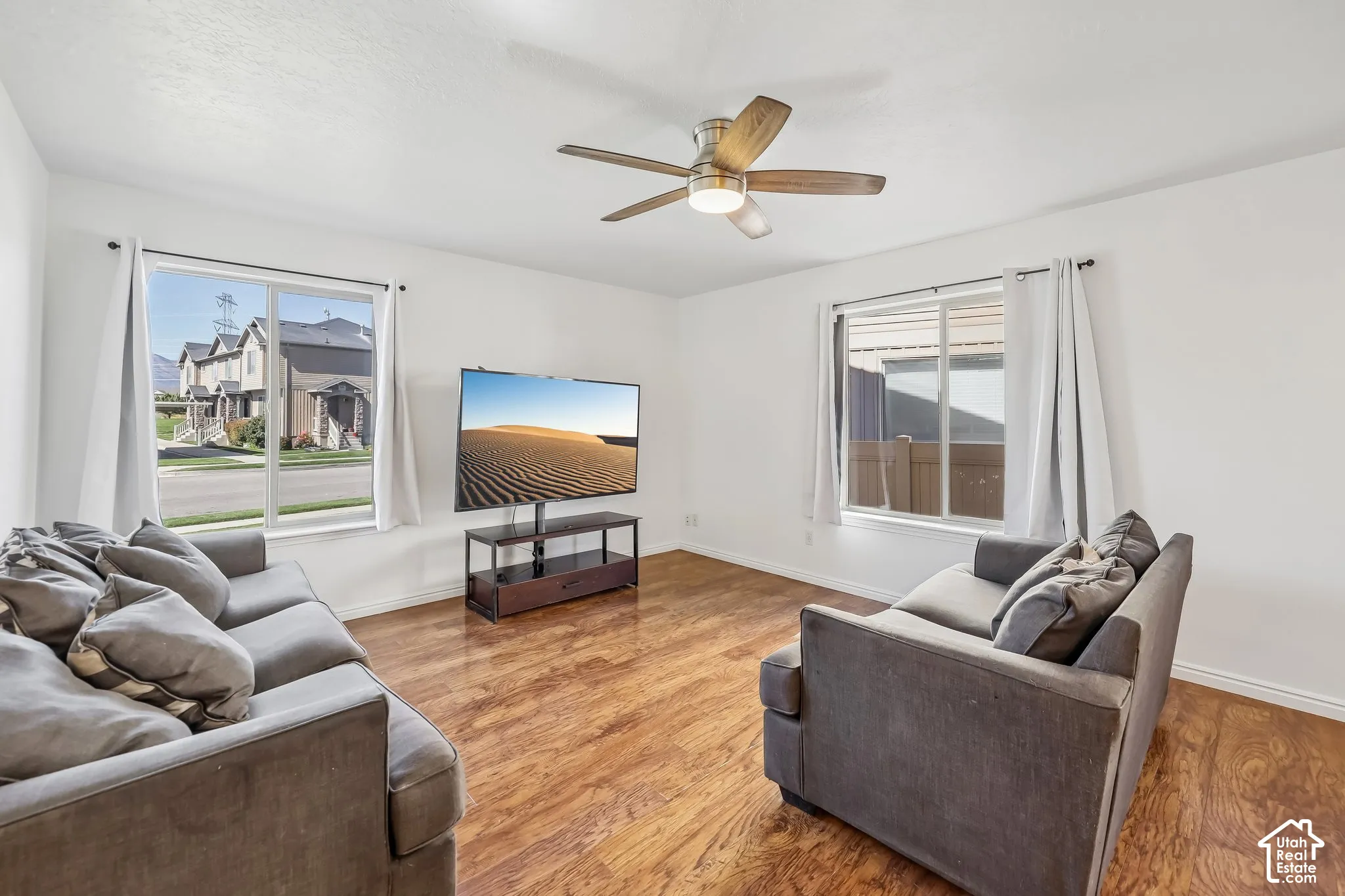 Spacious living area featuring wood-finish flooring, white walls, large windows, and a ceiling fan with integrated lighting