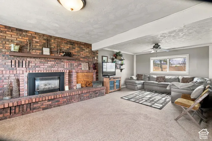 Spacious living area featuring a brick fireplace with a wood mantle, neutral carpeting, light gray walls, a ceiling fan, and a large window
