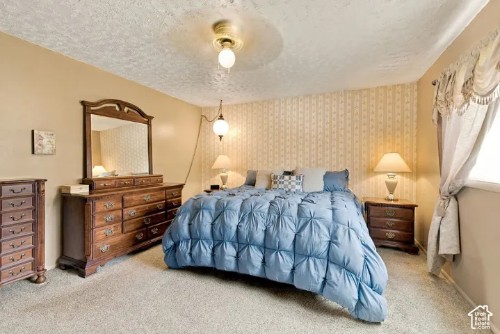 Carpeted flooring, textured ceiling, wall-mounted light fixture, wallpaper accent wall, window with valance