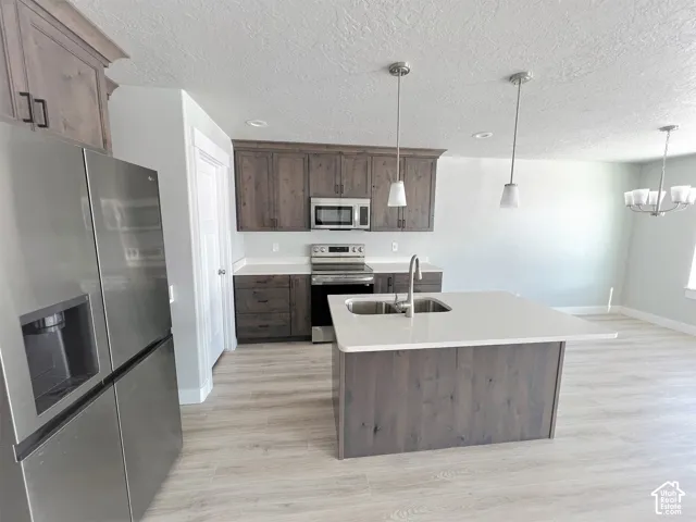 Kitchen featuring wood-finish cabinetry, stainless steel appliances, a center island with an undermount sink, pendant lighting, and wood-finish flooring