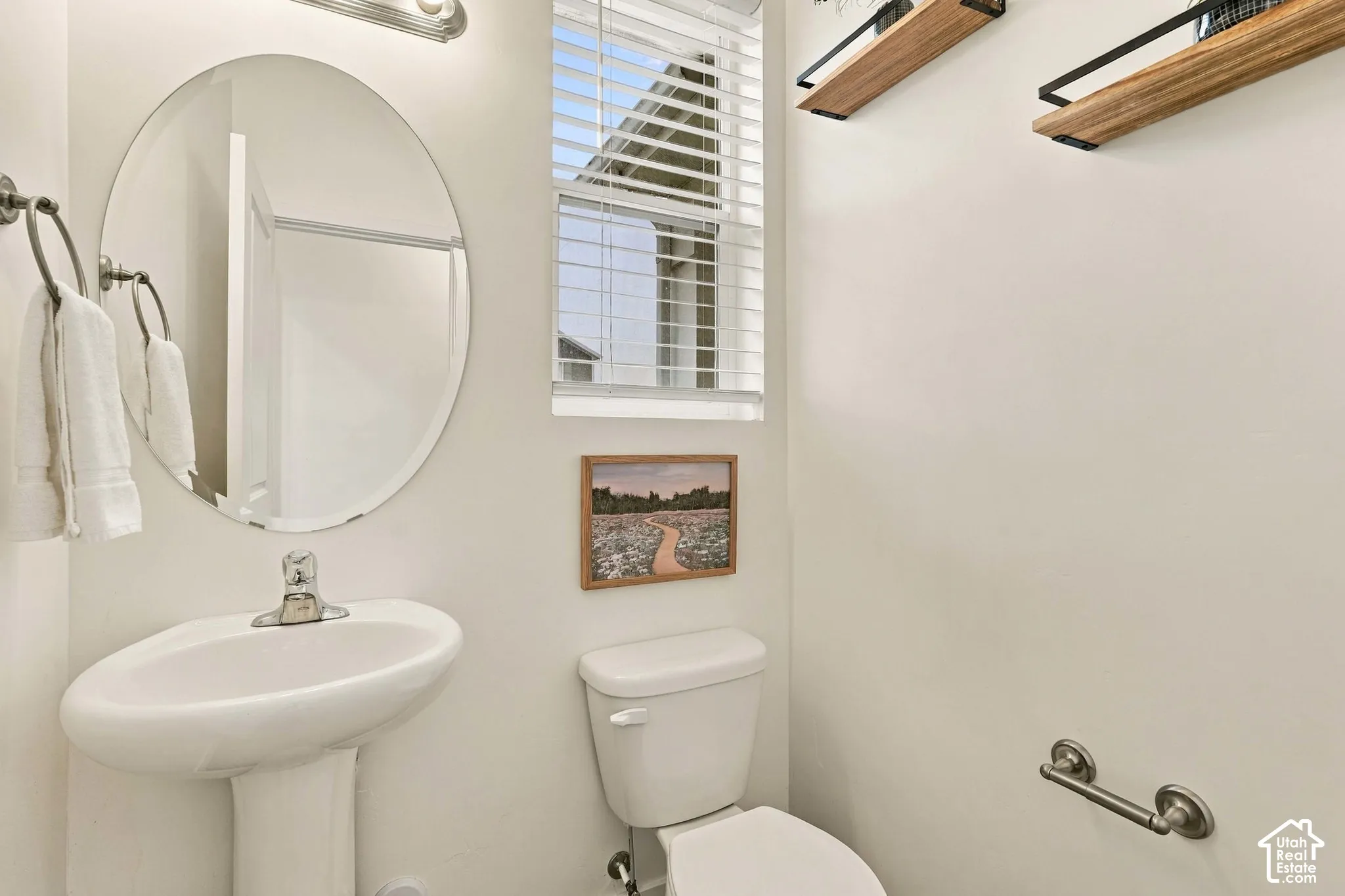 Pedestal sink with chrome faucet, oval mirror, window with blinds, and white toilet