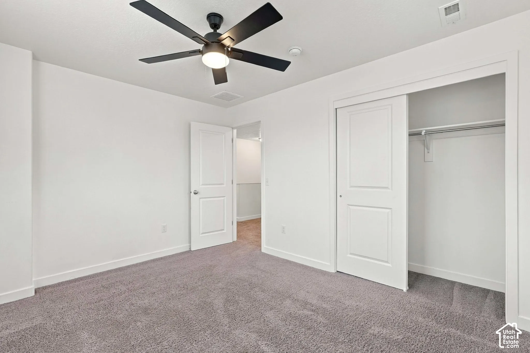 Spacious room featuring gray carpeting, white painted walls, a built-in closet with sliding doors, and a ceiling fan with integrated lighting