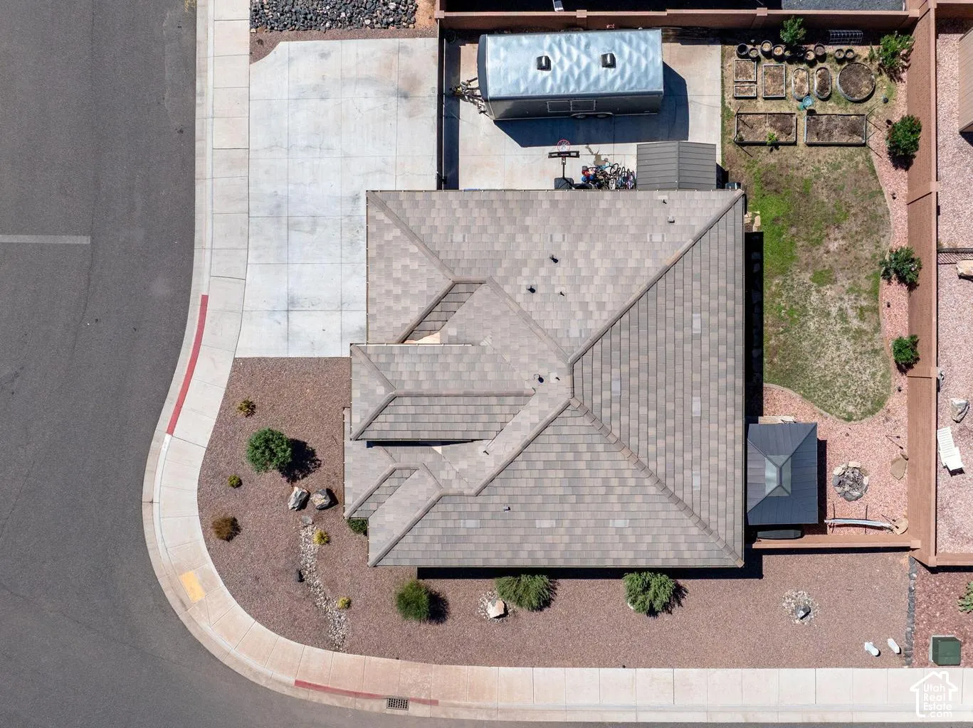 Pitched shingle roof with multiple gables, extensive concrete driveway, gravel landscaping with desert plantings, detached storage shed, and a private backyard with raised garden beds