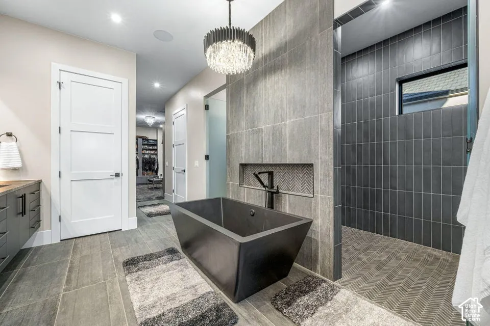 Porcelain tile flooring, freestanding soaking tub with matte black fixtures, tiled shower with chevron floor pattern, recessed shower niche, and a contemporary chandelier