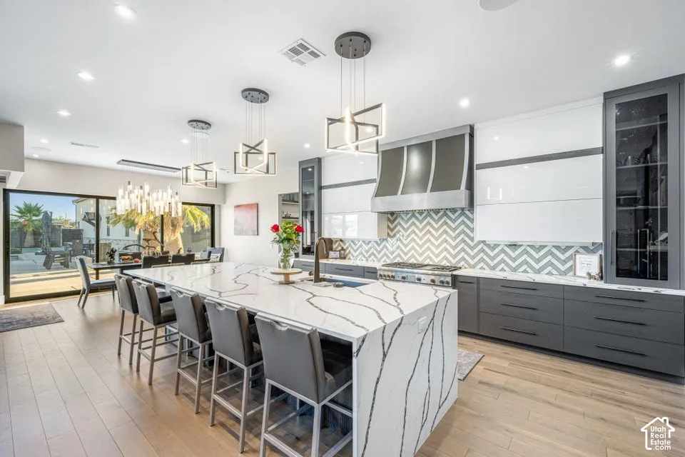 Spacious kitchen featuring a large waterfall island with integrated seating, a chevron tile backsplash, stainless steel range hood, and wood-finish flooring