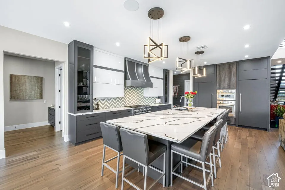Spacious kitchen featuring a large island with a waterfall edge, warm wood-finish flooring, custom cabinetry, a chevron-patterned backsplash, and integrated appliances