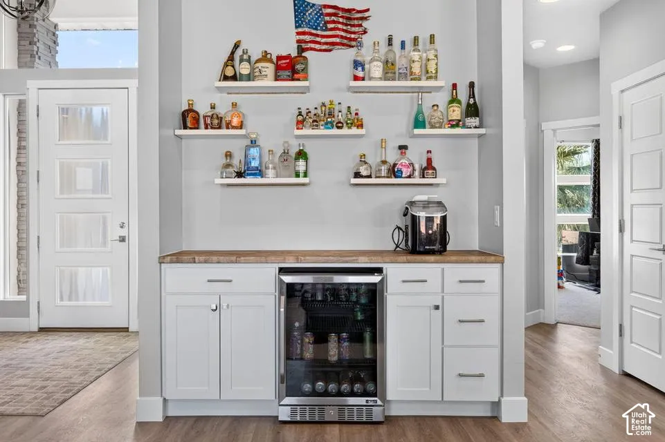 Built-in beverage station featuring a wood-finish countertop, white cabinetry with silver hardware, an integrated under-counter refrigerator, and wood-finish flooring