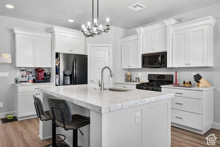 White shaker-style cabinetry, light-toned countertops, wood-finish flooring, central island with sink, and black appliances