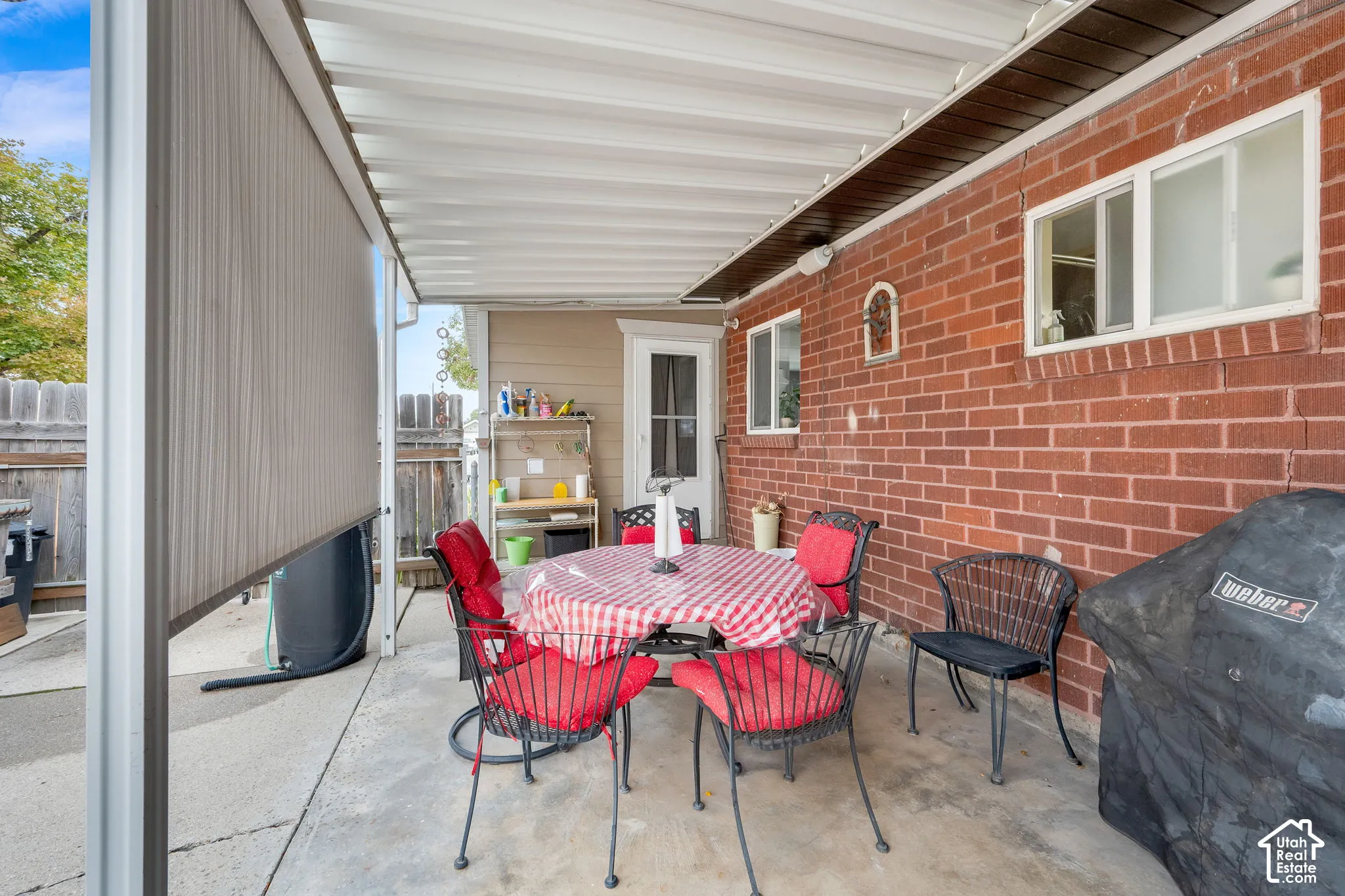 Covered concrete patio featuring a brick exterior wall, privacy shade, and white framed windows