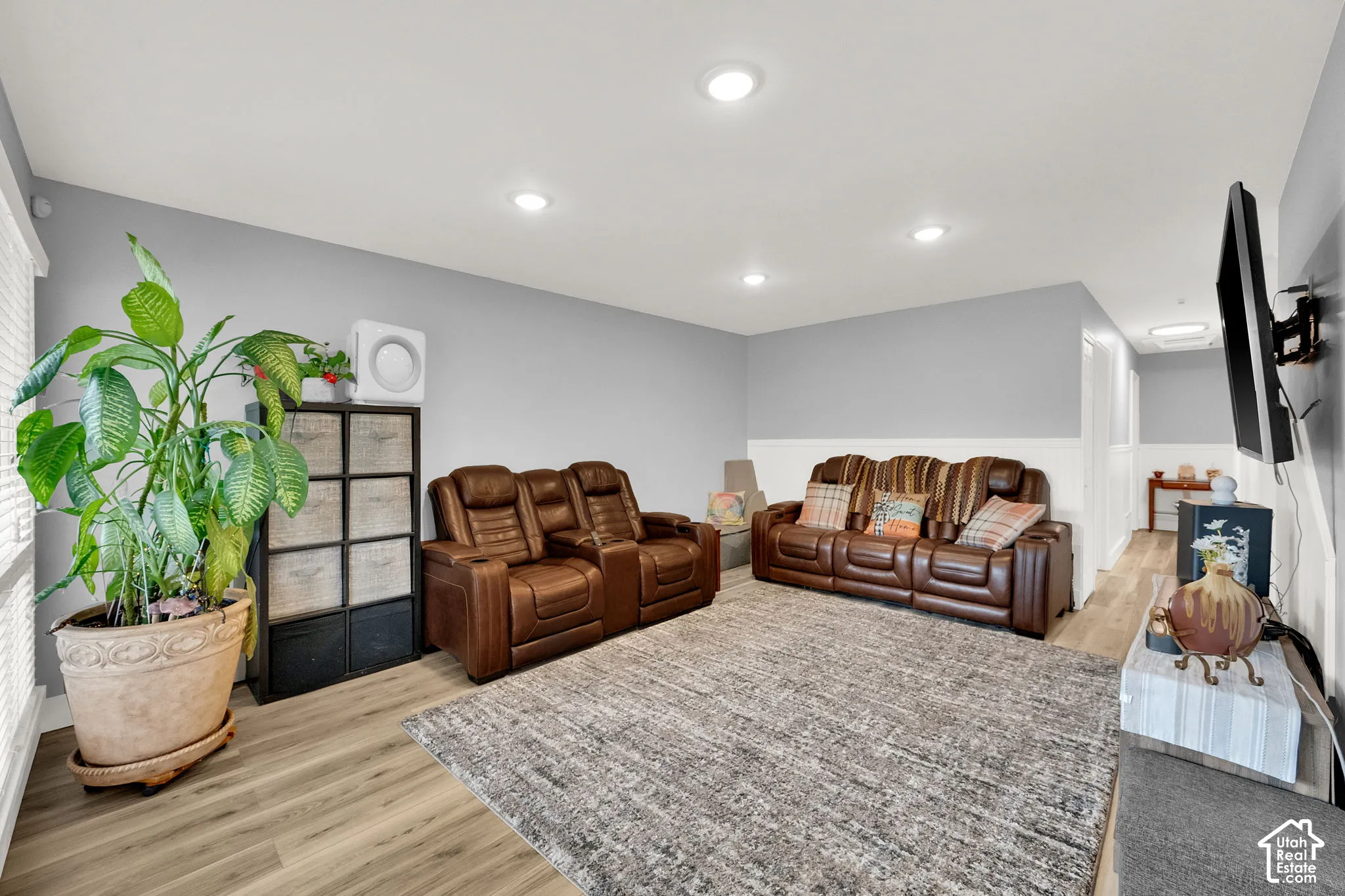 Recessed lighting illuminates the room featuring wood-finish flooring, a wall-mounted television bracket, and a half wall with white paneling
