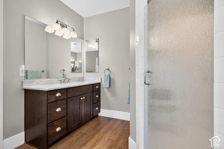 Wood-finish flooring, dark wood vanity with ample storage, white countertop, large wall-mounted mirror, and a textured glass shower enclosure
