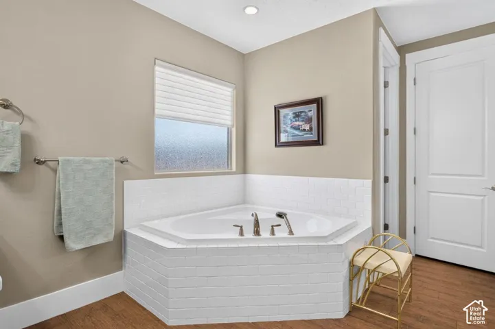 Corner soaking tub with white subway tile surround, wood-finish flooring, and a privacy window