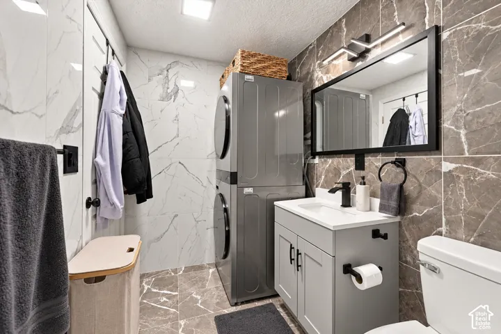 Floor-to-ceiling marbled tile, stacked laundry appliances, vanity with integrated sink, black framed mirror, and built-in toilet
