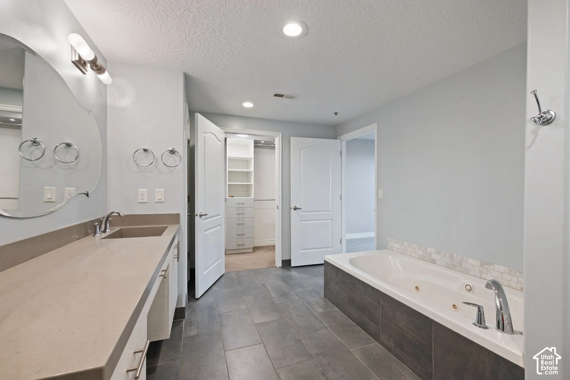 Double vanity with integrated sinks, a built-in jetted tub, gray tile flooring, and an adjoining closet with built-in shelving and drawers