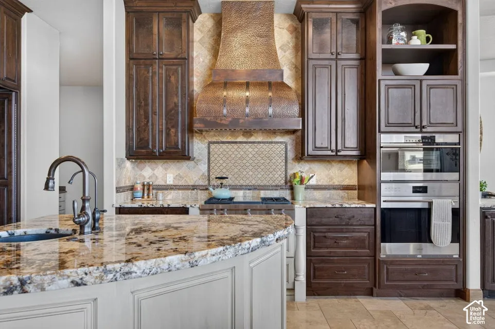 Kitchen featuring wood-finish cabinetry, built-in stainless steel appliances, a copper-tone range hood, a kitchen island with a built-in sink, and stone countertops