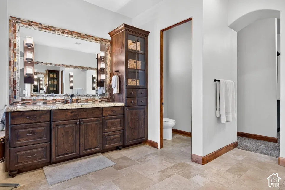 Dual vanity with rich wood cabinetry, integrated storage tower, expansive mirror with mosaic tile surround, and light-toned tile flooring