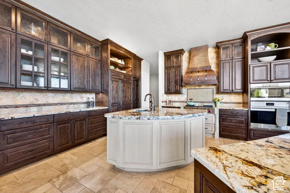 Kitchen featuring extensive dark wood cabinetry, a light-toned curved island with a sink, built-in stainless steel appliances, patterned backsplash, and light-toned tile flooring