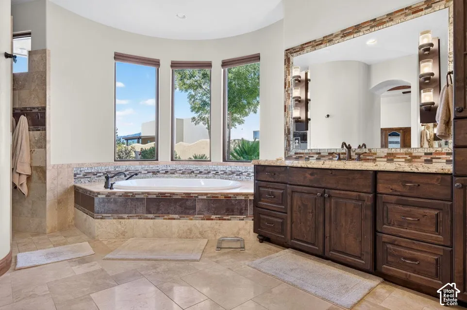 Soaking tub with mosaic tile surround, curved wall with three tall windows, large wood-finish vanity with stone countertop, and an oversized mirror with decorative frame