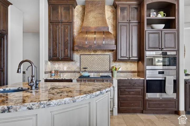 Kitchen featuring wood-finish cabinetry, built-in stainless steel appliances, a copper-tone range hood, a kitchen island with a built-in sink, and stone countertops