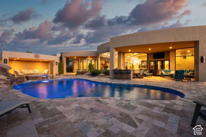 Curved-edge swimming pool with integrated waterfall feature, expansive patio with stone-finish pavers, recessed covered patio with built-in bar, additional covered patio with built-in ceiling lights, and large floor-to-ceiling windows