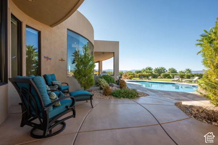 Curved architectural exterior with large windows, expansive concrete patio, built-in swimming pool, and landscaping