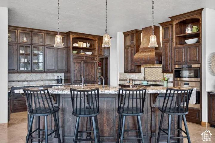 Kitchen featuring dark wood cabinetry, a large island with a patterned countertop, three pendant lights, a mosaic tile backsplash, and stainless steel appliances