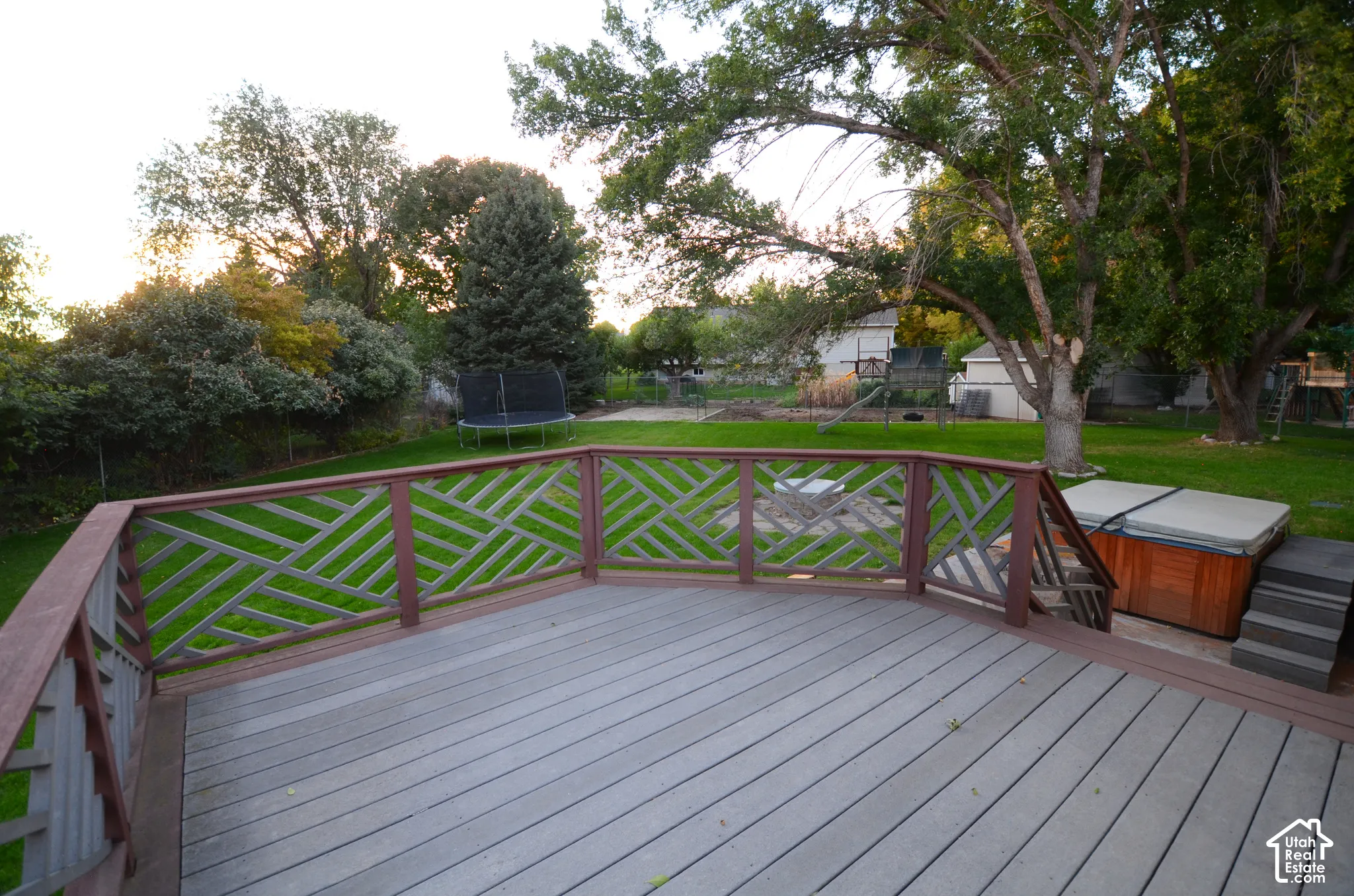 Expansive composite deck with decorative railing, built-in hot tub, and a large grassy yard featuring mature trees
