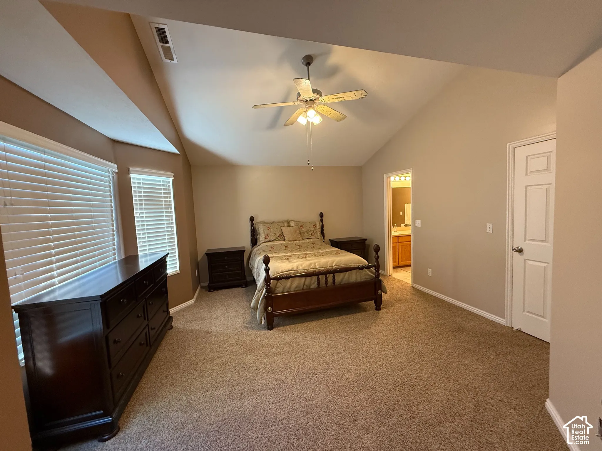 Vaulted ceiling, ceiling fan, multiple windows with blinds, carpeted flooring, and an adjoining bathroom with a vanity and mirror