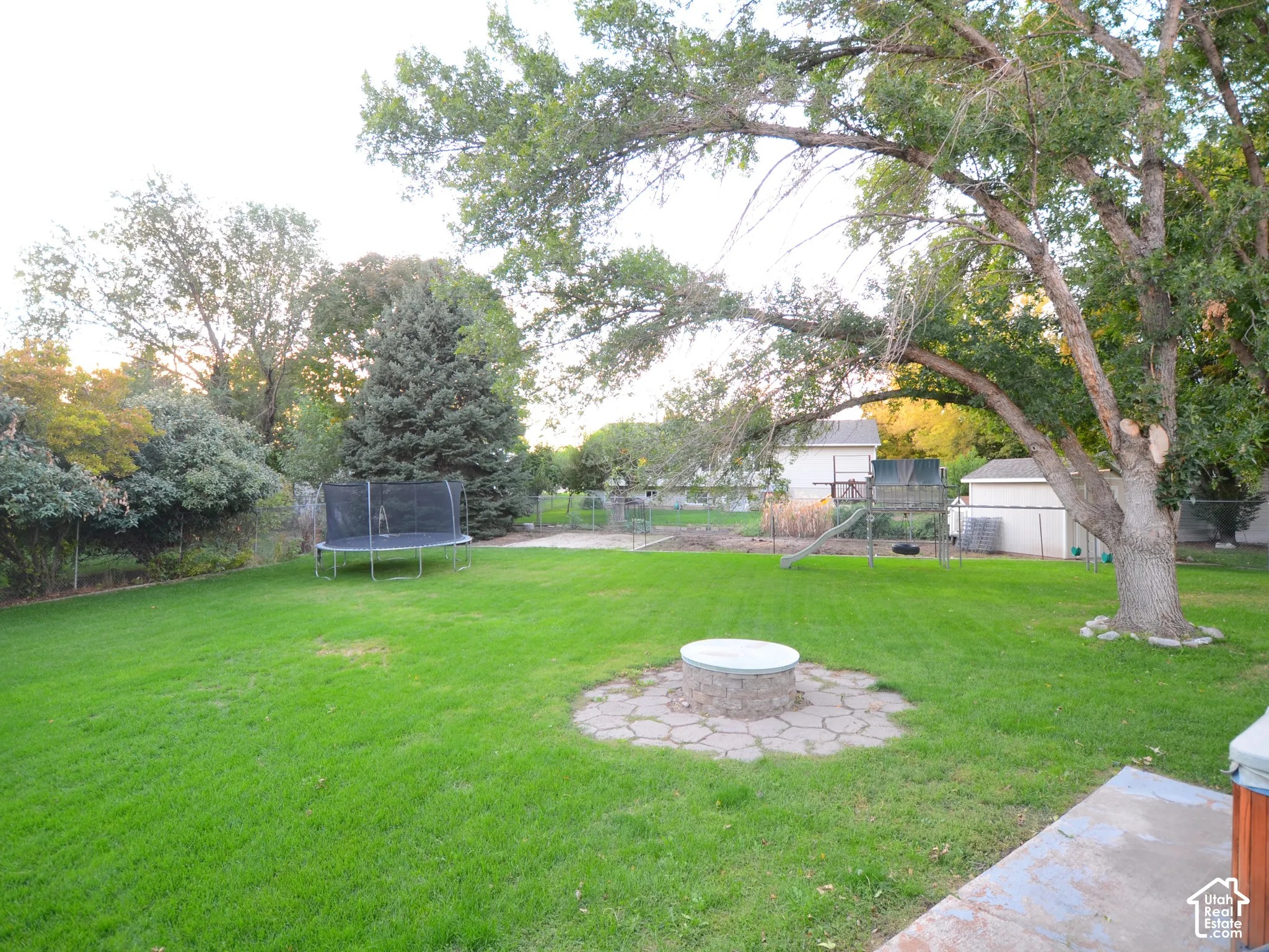 Expansive backyard oasis featuring a manicured lawn, mature shade trees, and a stone-lined fire pit