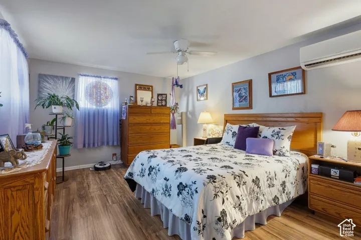 Wood-finish flooring with a ceiling fan, wall-mounted air conditioning unit, and a window with sheer coverings