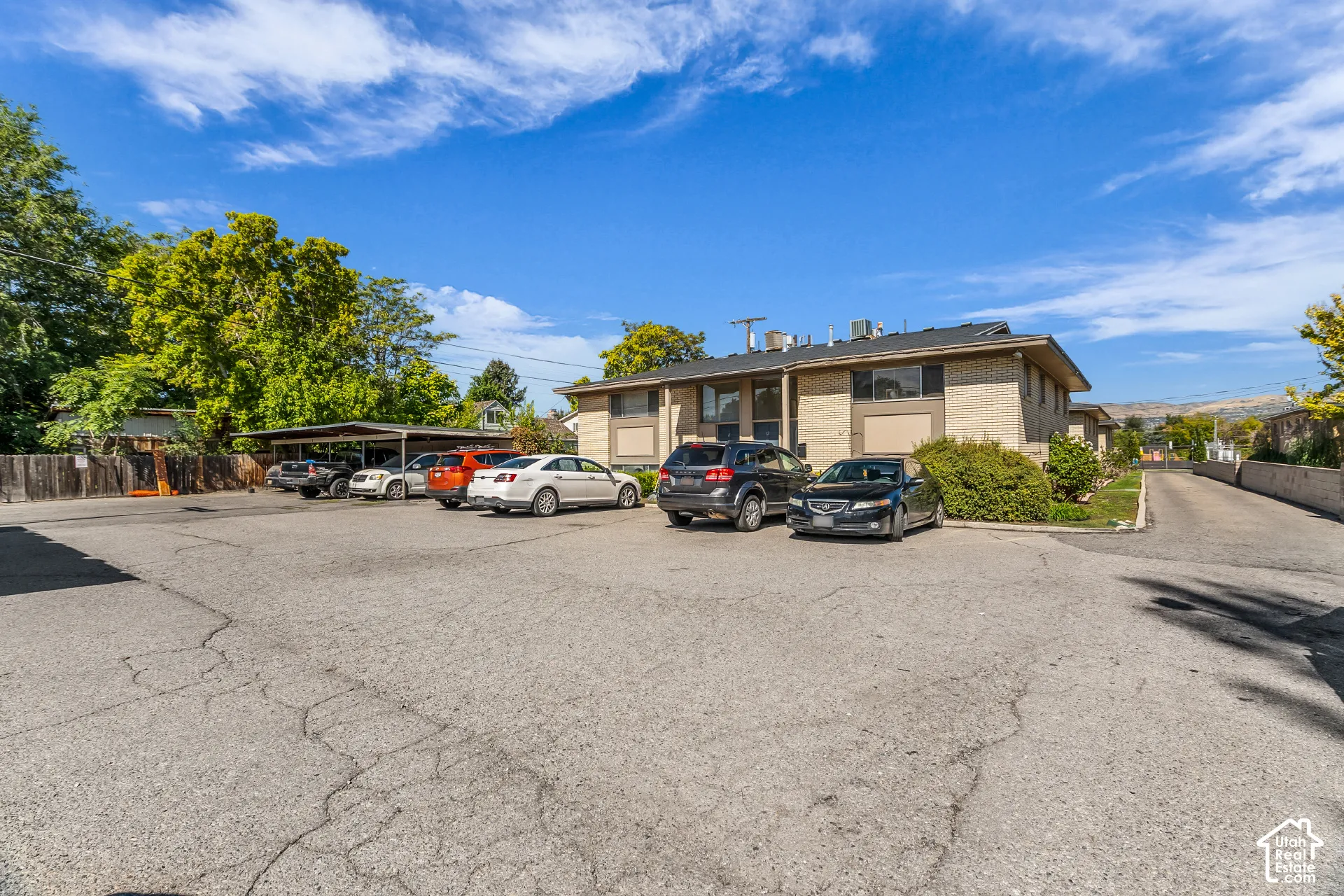 Paved parking lot with designated covered parking area, brick exterior building with dark window frames, and surrounding mature trees