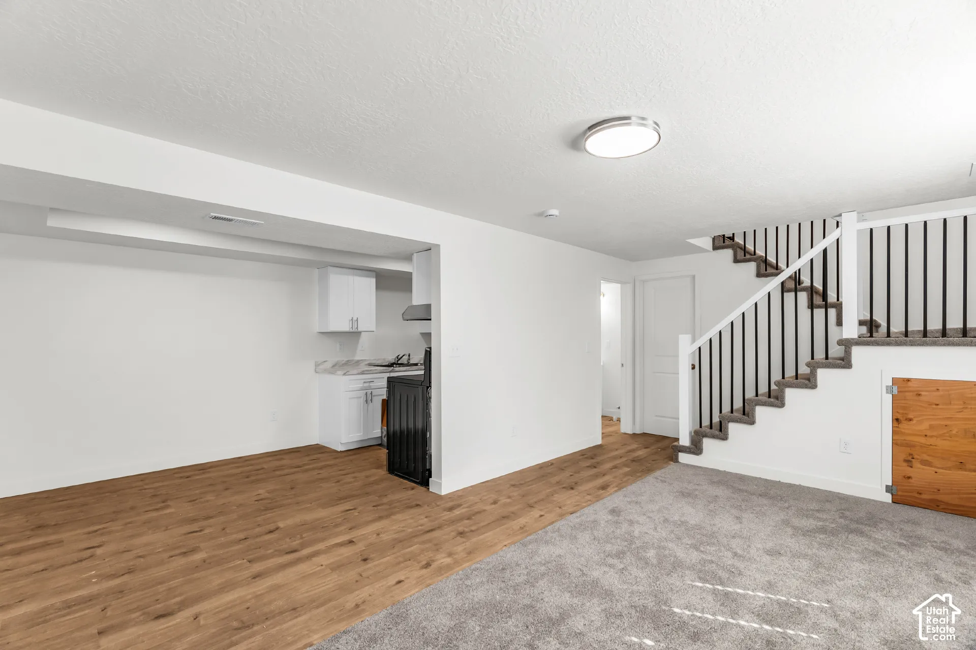 Open-concept room featuring wood-finish flooring, a kitchen area with white cabinetry, a built-in oven, and a staircase with a white railing and dark balusters