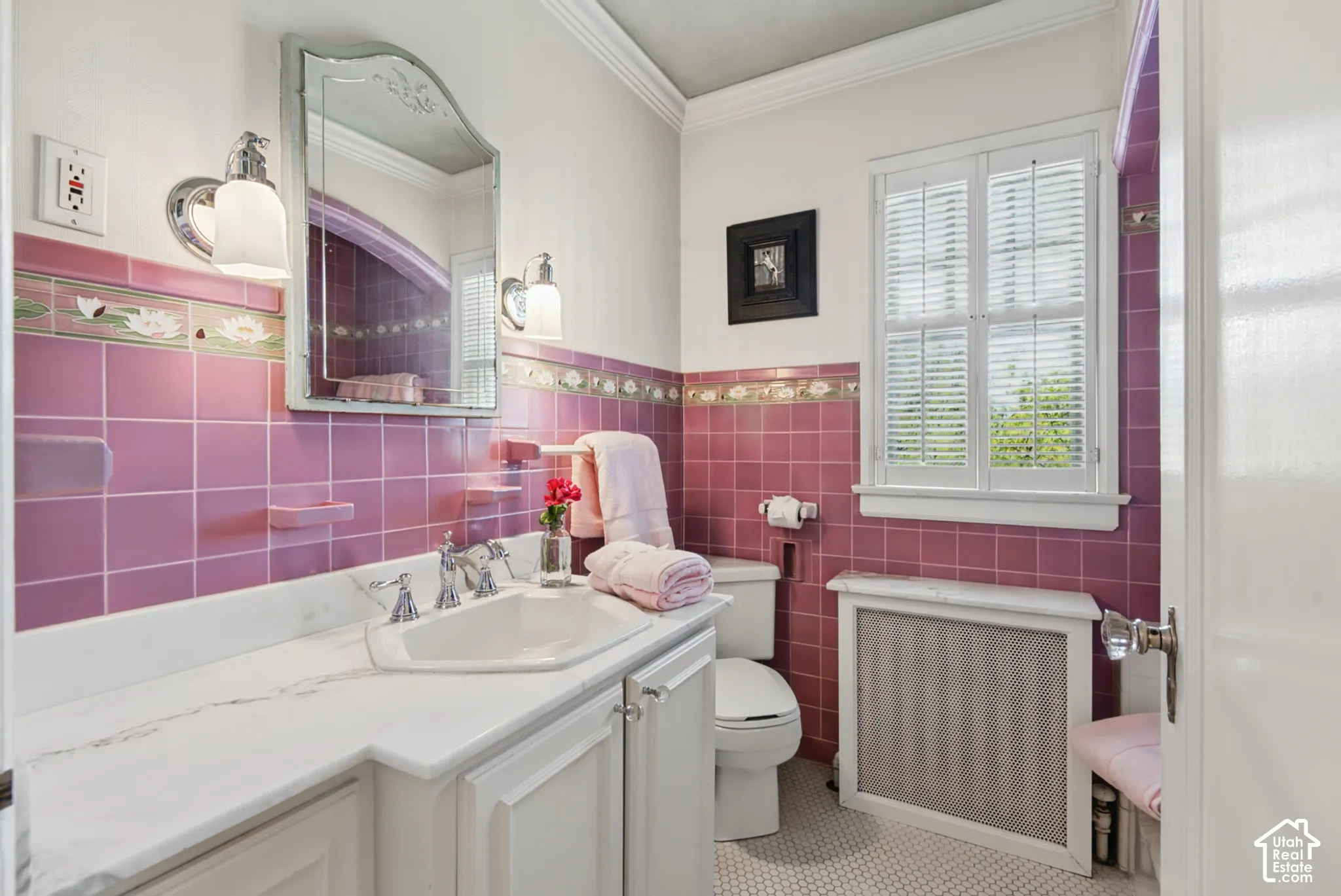 Bathroom featuring pink tiled walls, a window with louvered shutters, white vanity cabinetry with a built-in sink, a wall-mounted mirror, and a radiator cover