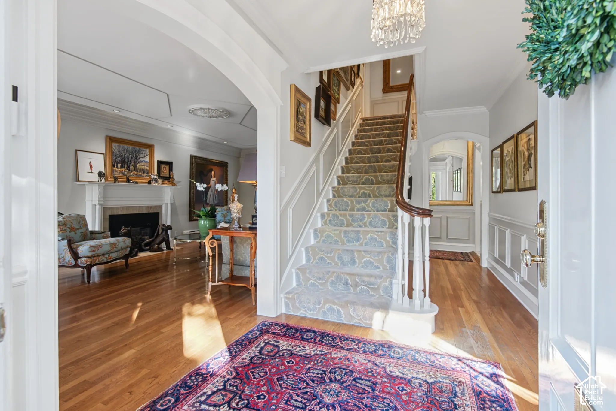 Hardwood flooring, arched doorway, grand staircase with decorative runner, wainscoting, and an ornate chandelier