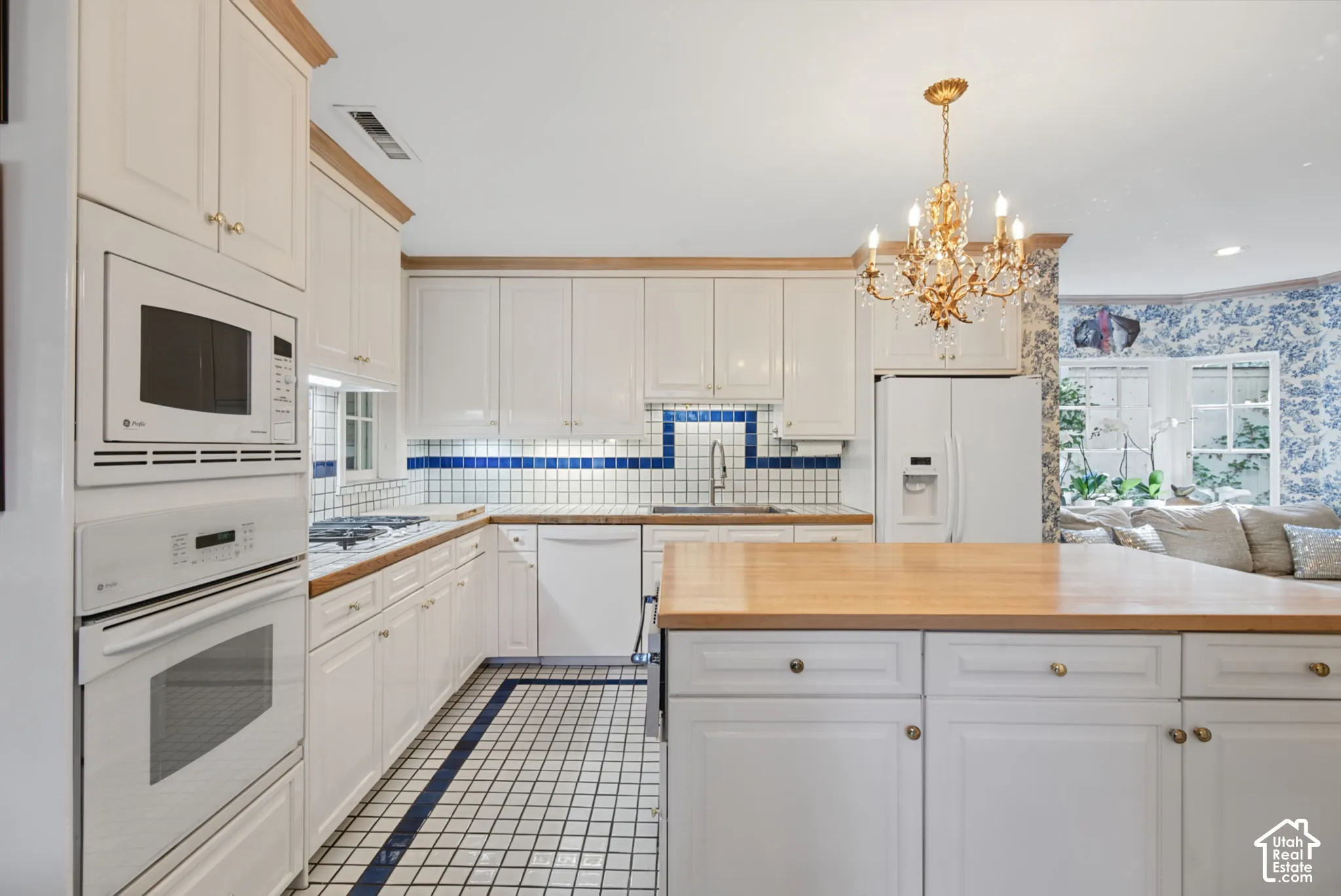 White cabinetry with wood trim, integrated white appliances, blue and white tiled backsplash, gold-toned chandelier, and a wood-finish island countertop