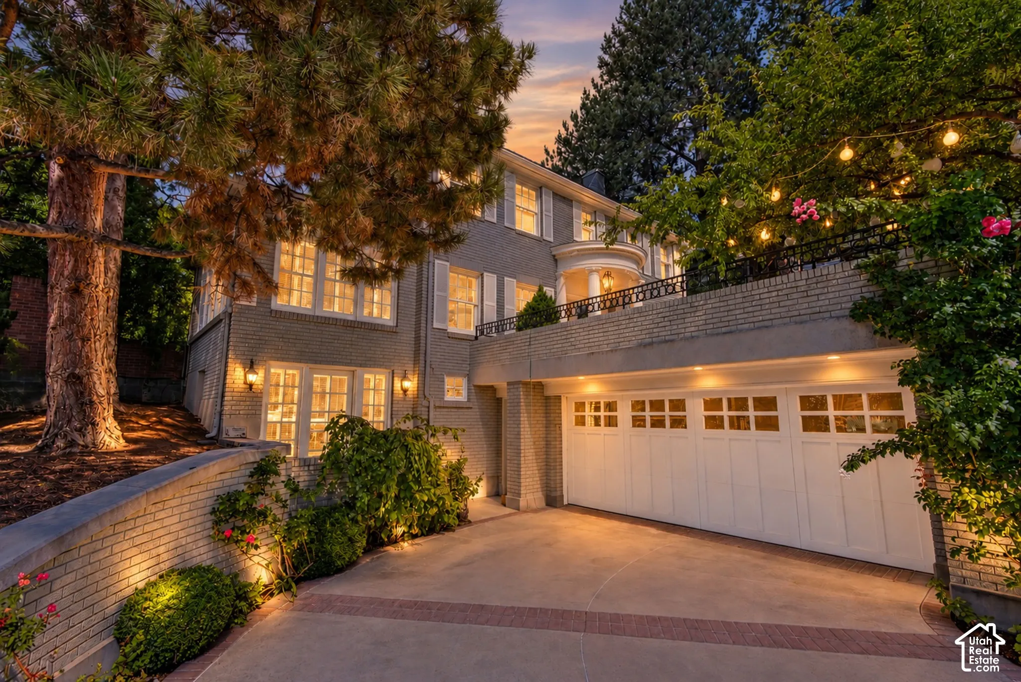 Brick exterior with white window shutters and trim, featuring a recessed garage with multiple doors, a prominent balcony, and mature trees