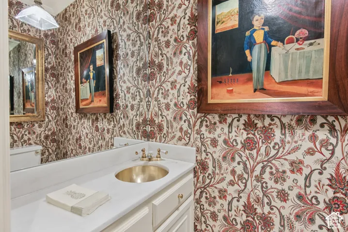 Patterned wallpaper, white vanity with integrated sink, brass fixtures, gold-framed mirror, and surface-mounted ceiling light fixture