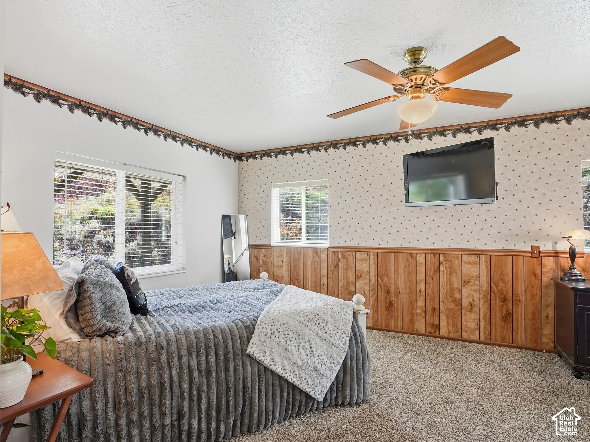 Room featuring wood-panel wainscoting, decorative wallpaper with a border, wall-to-wall carpeting, multiple windows with blinds, and a ceiling fan with integrated light fixture