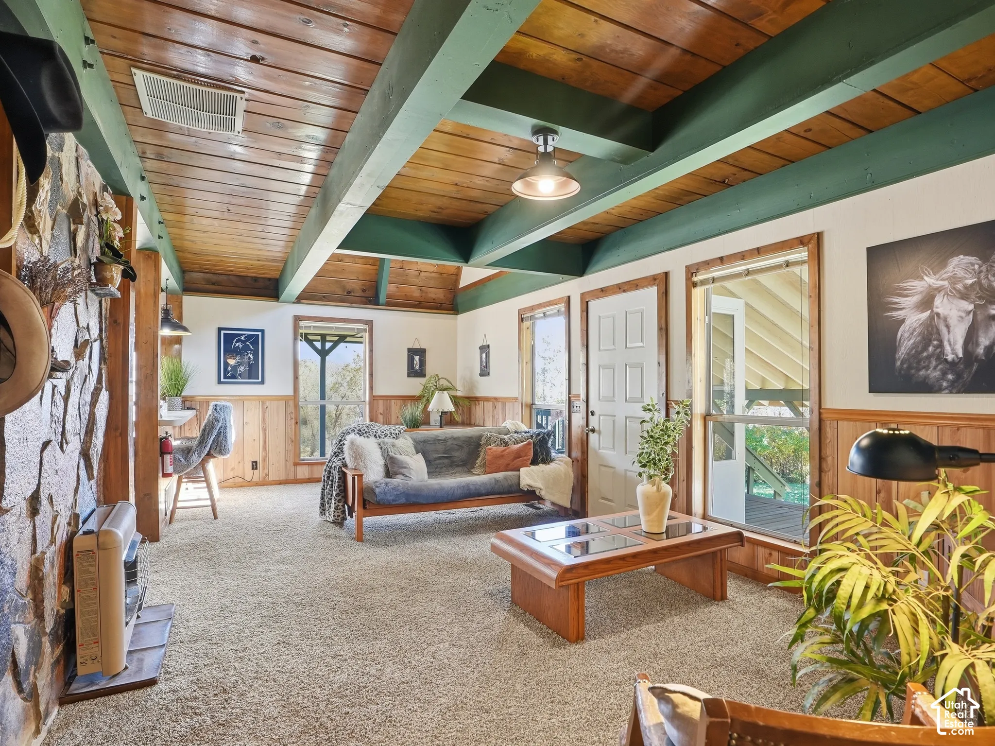 Exposed wood plank ceiling with structural beams, stacked stone fireplace, wood-paneling wainscoting, and multiple exterior access points