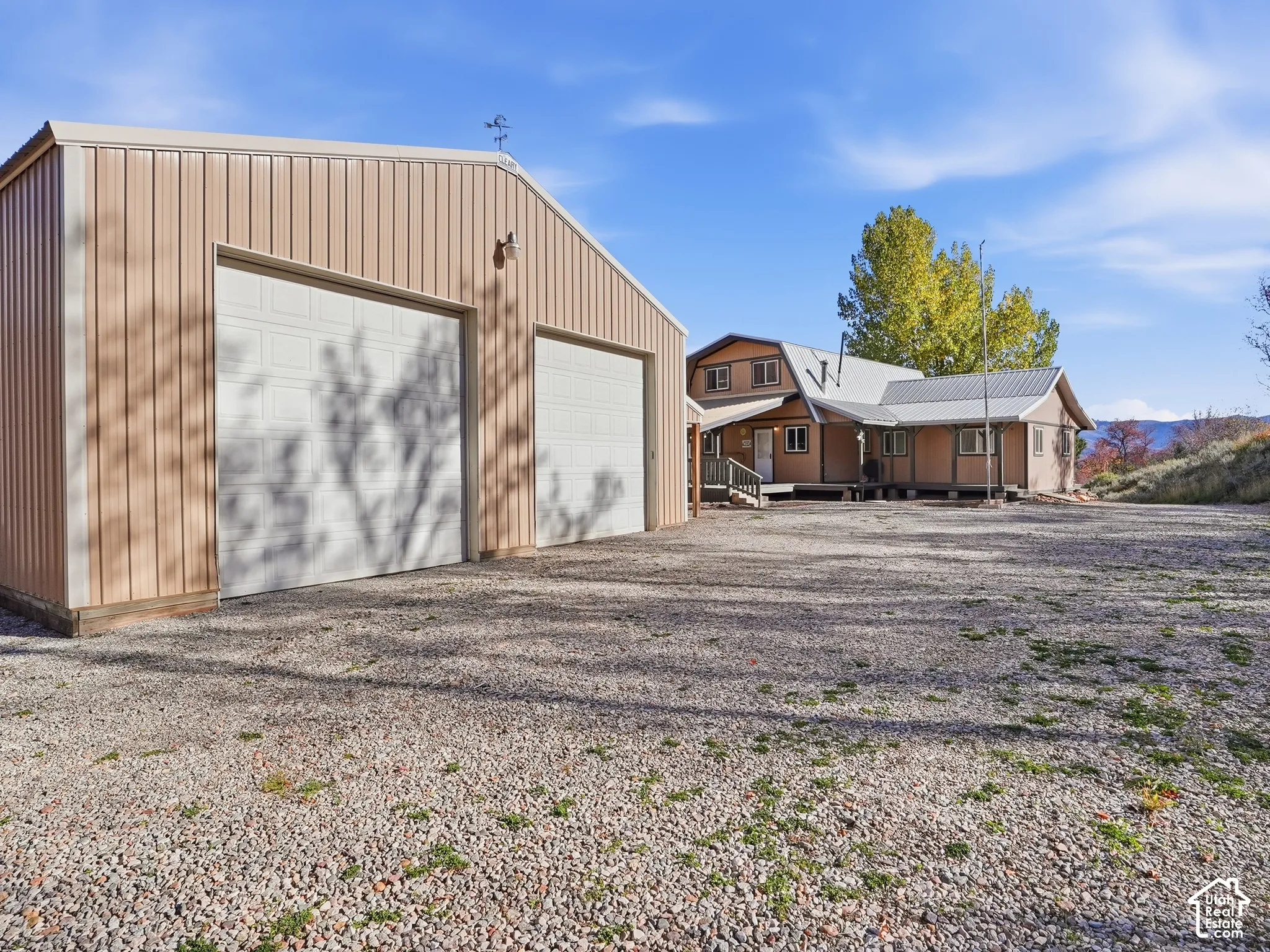 Detached metal-sided garage with two roll-up doors, gravel drive, and multi-story residence with a metal roof