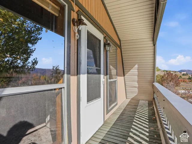 Covered exterior balcony featuring horizontal slat flooring, vertical siding, wall-mounted light fixtures, and a glass-paneled screen door